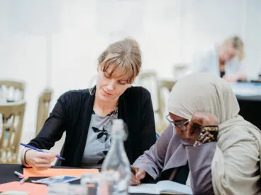 Two people writing at a table amongst other tables of people